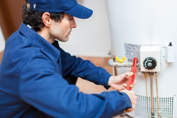 What Are the Different Types of Water Heaters? Image is a photograph of an HVAC professional in a blue jumpsuit and baseball cap working on a water heater.