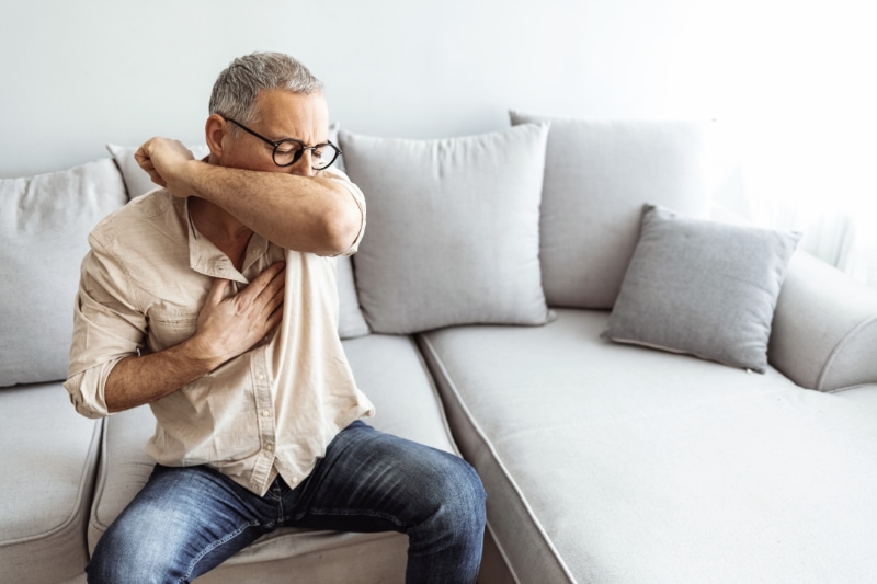 How to Spot a Cracked Heat Exchanger. Caucasian male coughs in his elbow while sitting on sofa at home. Correct sneezing. Concept of stop spread of the virus.