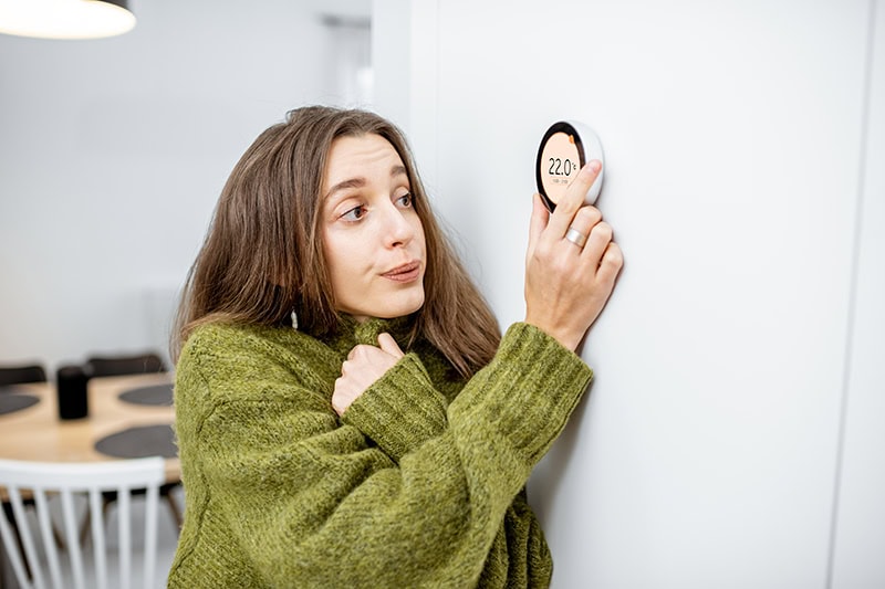 woman shivering in a sweater while she turns up her thermostat.