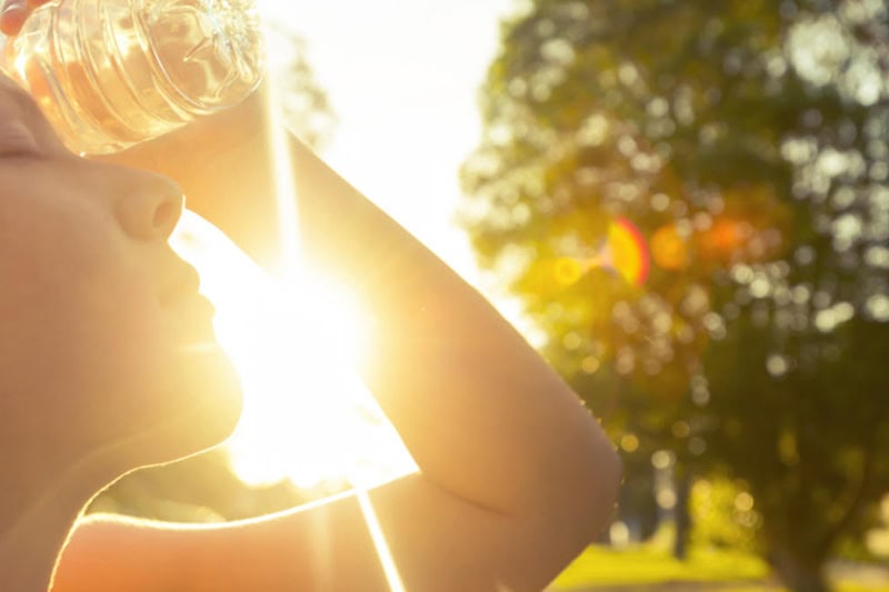 Why Is My AC Blowing Hot Air? Photo of a woman trying to cool off with a water bottle in the summer sun.